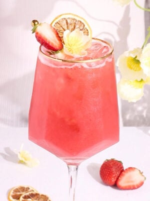 A pink mocktail with ice in a stemmed glass, garnished with a lemon slice, half a strawberry, and a yellow flower. Fresh strawberries and dried lemon slices are on the white table nearby.