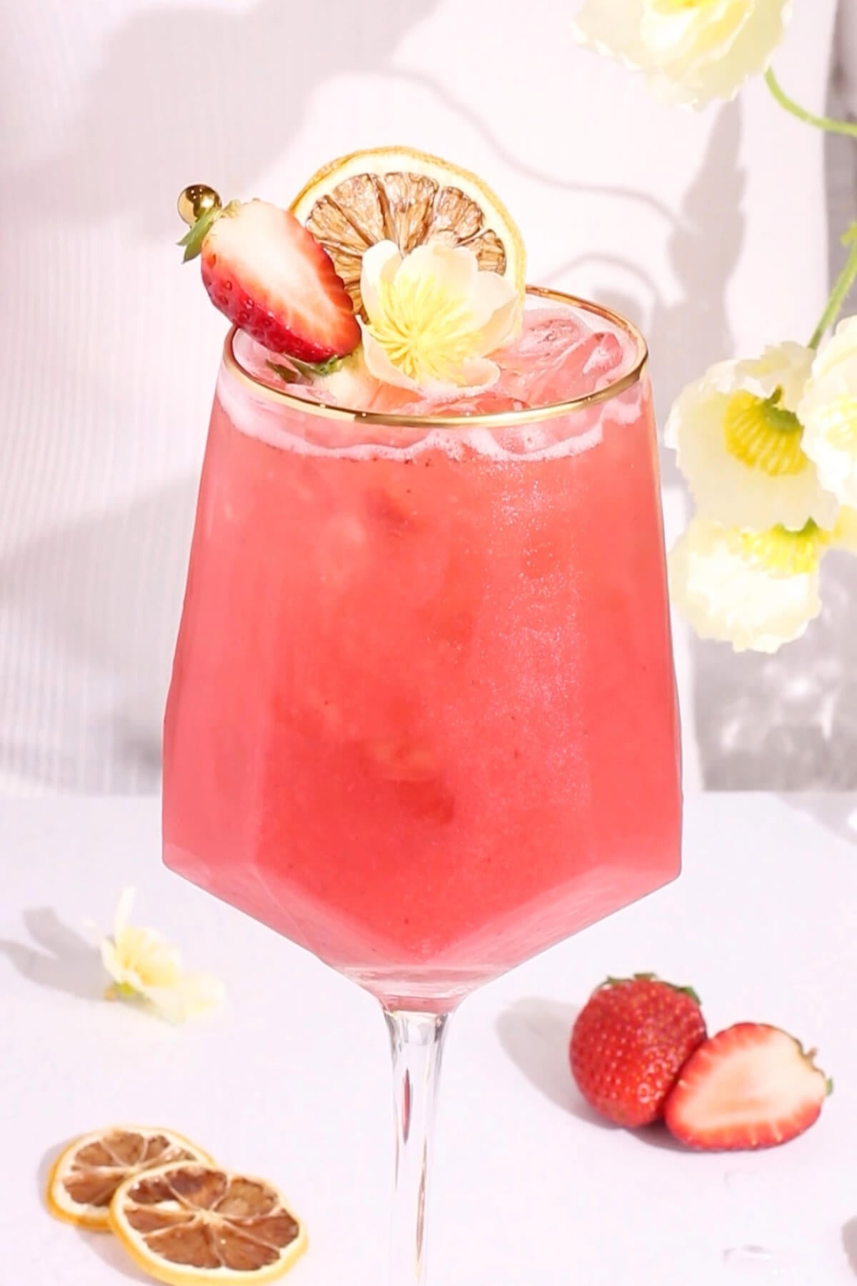 A pink mocktail with ice in a stemmed glass, garnished with a lemon slice, half a strawberry, and a yellow flower. Fresh strawberries and dried lemon slices are on the white table nearby.