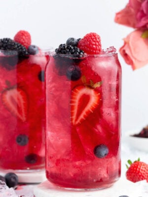 Two clear glasses filled with bright red berry punch, ice, and garnished with strawberries, blueberries, blackberries, and raspberries; a pink flower bouquet and scattered berries are in the background.