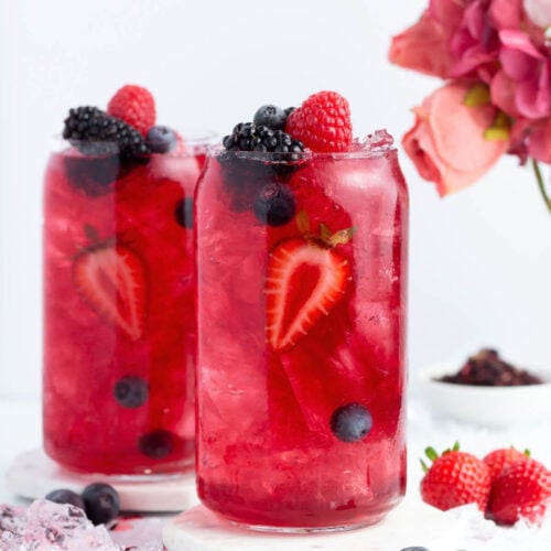 Two clear glasses filled with bright red berry punch, ice, and garnished with strawberries, blueberries, blackberries, and raspberries; a pink flower bouquet and scattered berries are in the background.