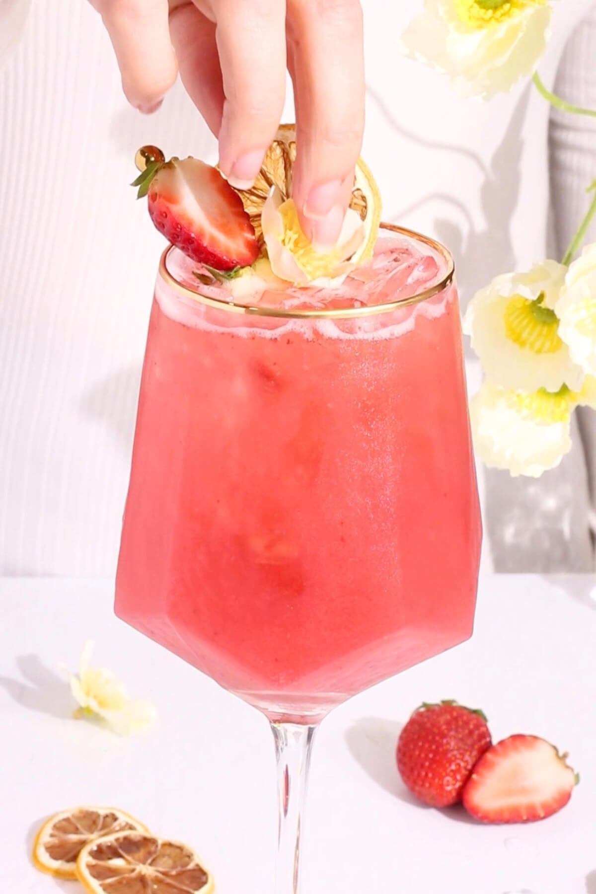 A hand garnishes a vibrant pink drink in a stemmed glass with a slice of lemon and a strawberry. Fresh strawberries and dried citrus slices sit on the white surface nearby. Yellow flowers are visible in the background.