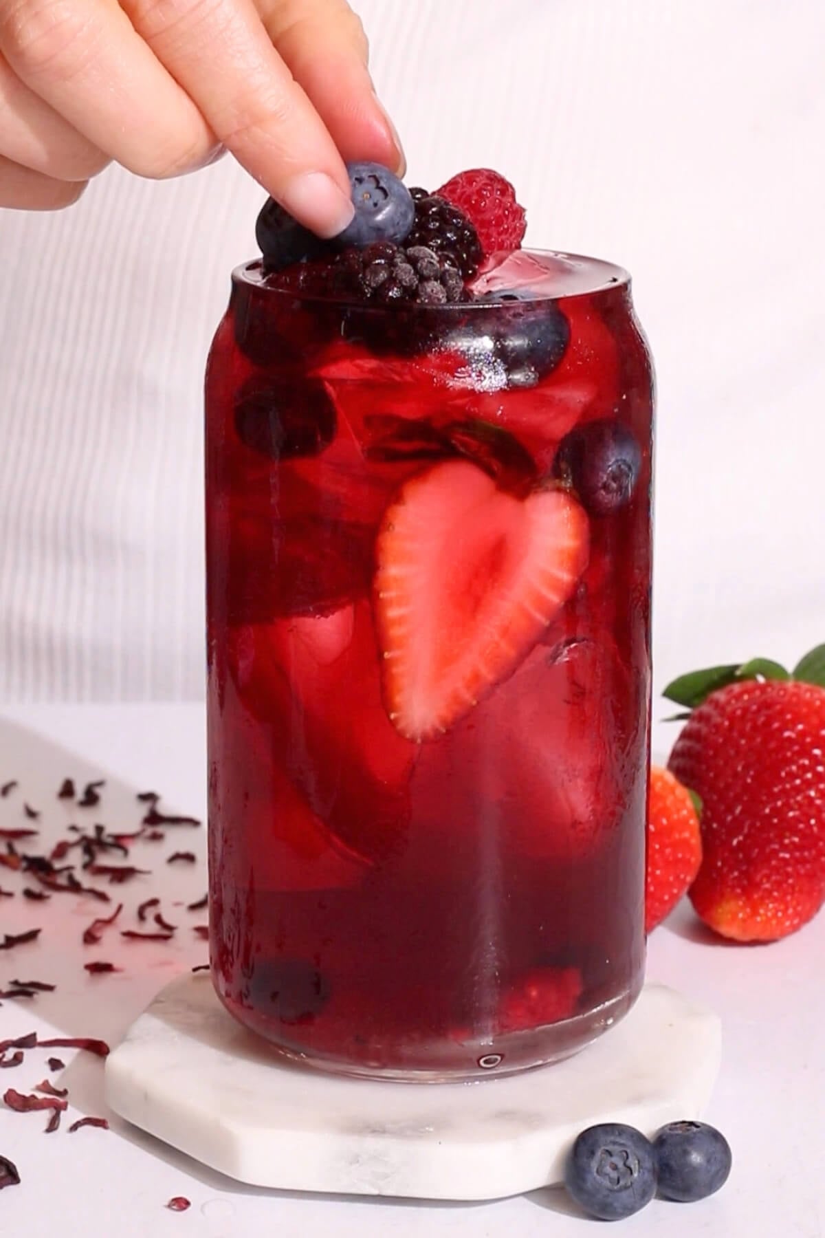 A hand is placing blueberries on top of a glass filled with iced red berry drink, garnished with sliced strawberries and assorted berries. Whole strawberries and blueberries are on the white surface nearby.