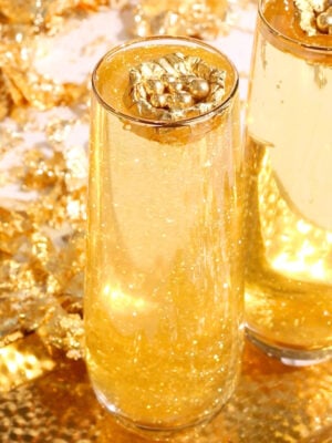Two tall glasses filled with sparkling golden liquid topped with decorative gold leaf accents, set against a background of gold foil and shimmering gold decorations.