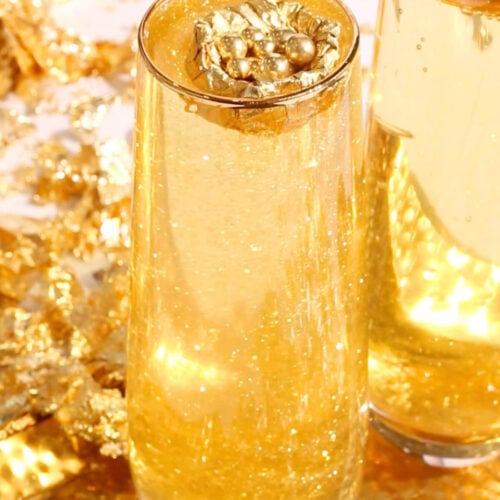 Two tall glasses filled with sparkling golden liquid topped with decorative gold leaf accents, set against a background of gold foil and shimmering gold decorations.