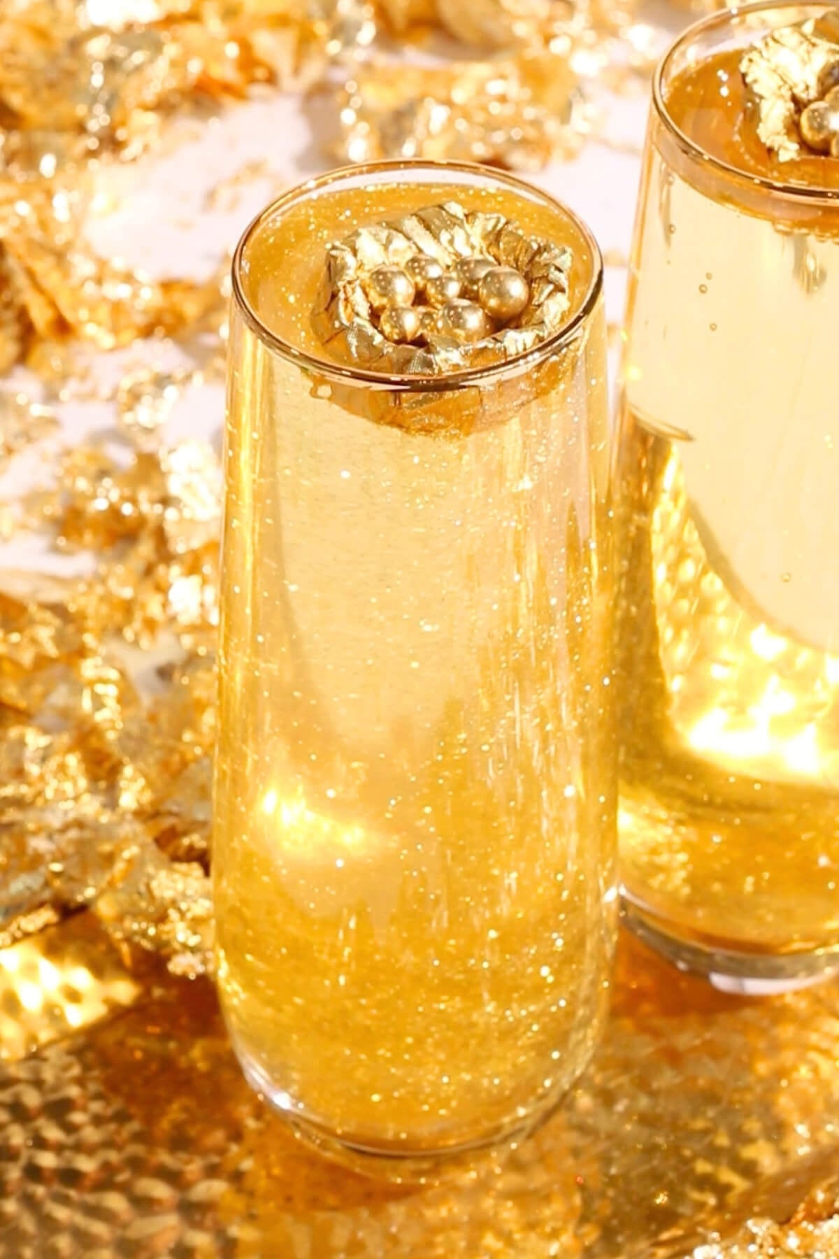Two tall glasses filled with sparkling golden liquid topped with decorative gold leaf accents, set against a background of gold foil and shimmering gold decorations.
