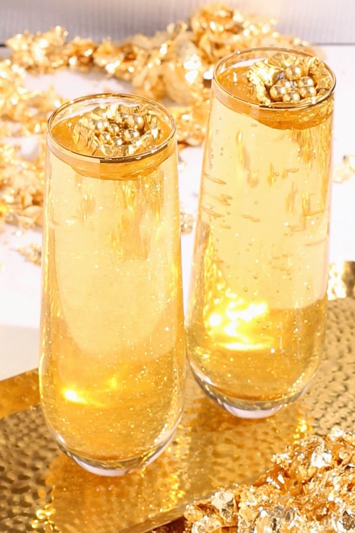 Two tall glasses filled with sparkling golden mocktails, each garnished with a cluster of shiny gold decorations. Gold sequins and metallic accents surround the glasses, creating a festive, celebratory atmosphere.