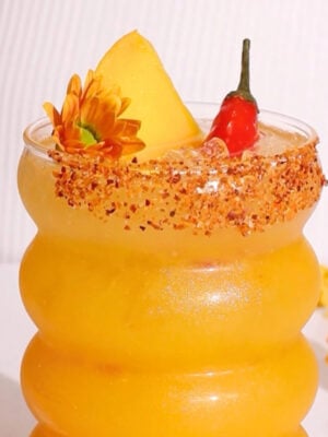 A vibrant yellow drink in a curvy glass, garnished with a mango slice, small red chili, and orange flowers. The rim is coated with chili powder, and the background is white with a few flowers beside the glass.
