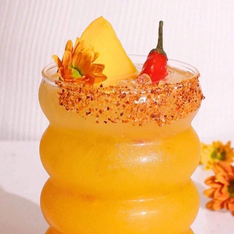 A vibrant yellow drink in a curvy glass, garnished with a mango slice, small red chili, and orange flowers. The rim is coated with chili powder, and the background is white with a few flowers beside the glass.