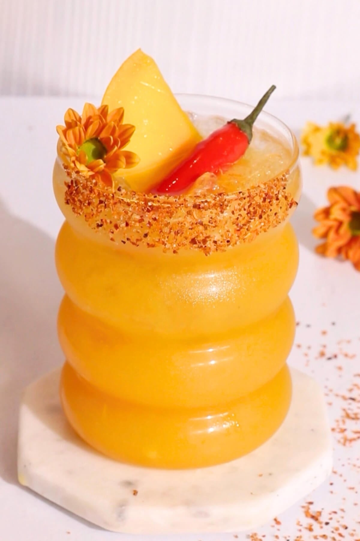 A vibrant orange cocktail in a wavy glass, topped with a mango slice, red chili, and orange flowers. The glass rim is coated with chili powder, and it sits on a round marble coaster.