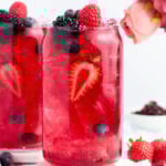 A tall glass filled with a bright red berry drink, ice, and garnished with sliced strawberries, raspberries, blackberries, and blueberries. A bouquet of pink flowers and more berries are in the background.