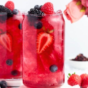 A tall glass filled with a bright red berry drink, ice, and garnished with sliced strawberries, raspberries, blackberries, and blueberries. A bouquet of pink flowers and more berries are in the background.