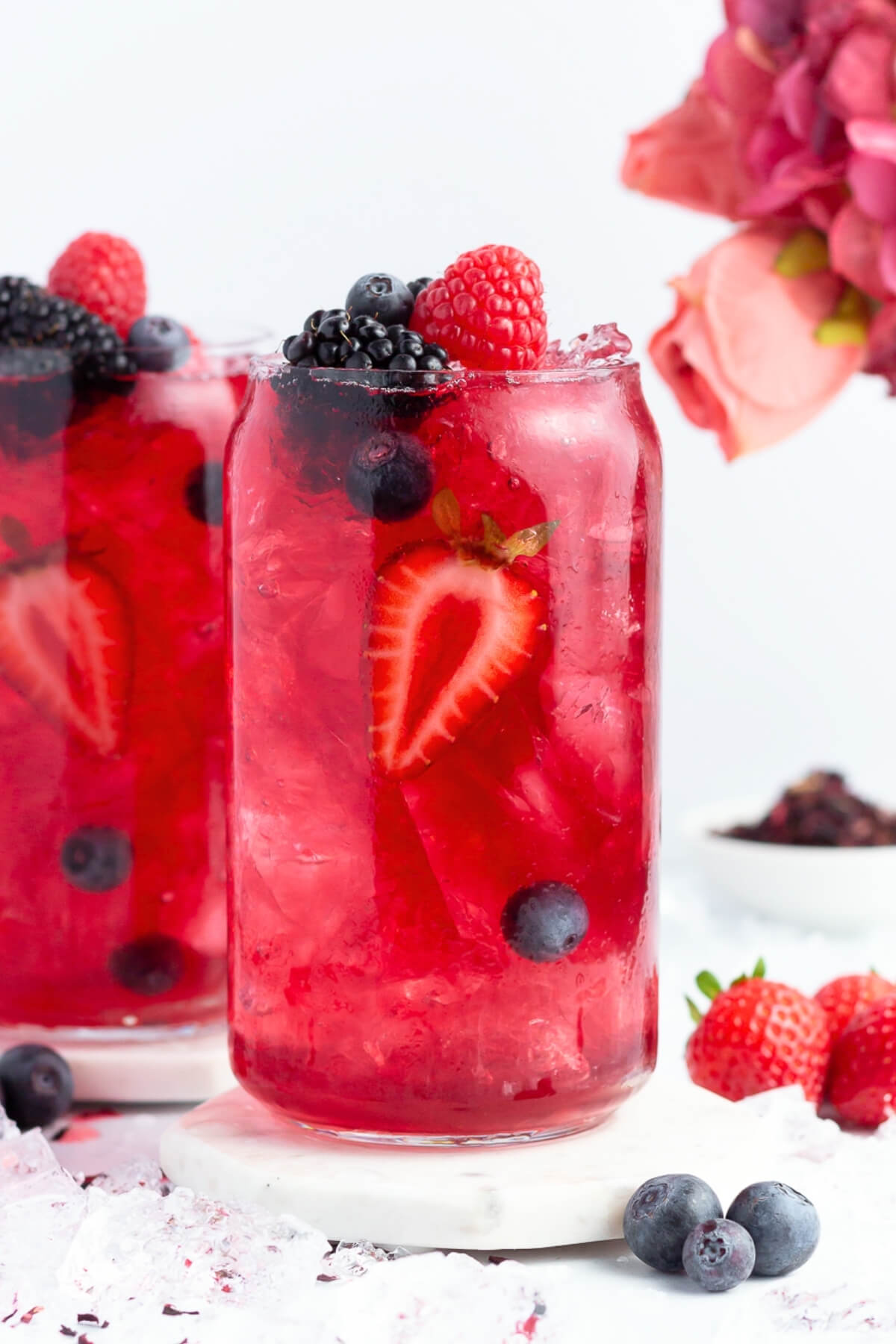 A tall glass filled with a bright red berry drink, ice, and garnished with sliced strawberries, raspberries, blackberries, and blueberries. A bouquet of pink flowers and more berries are in the background.