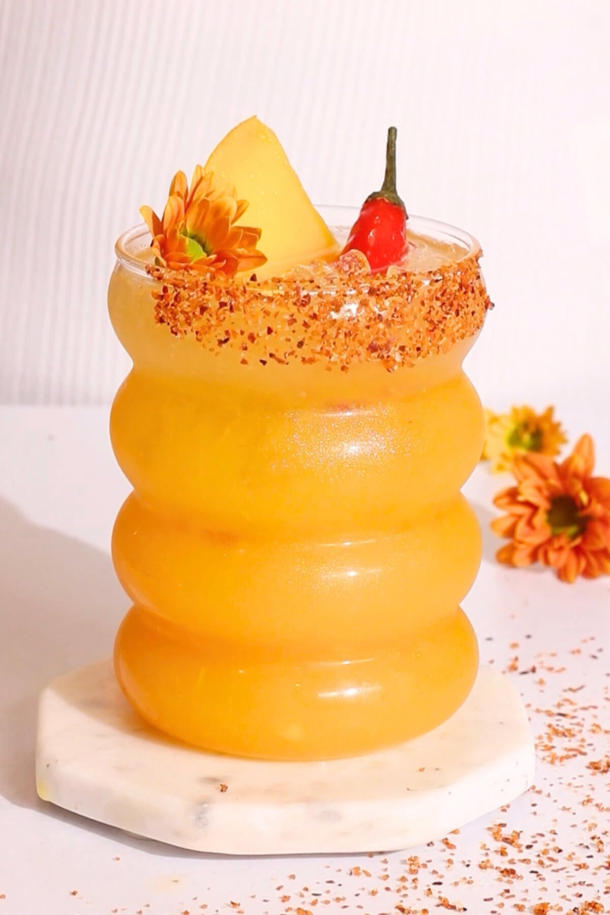 A vibrant orange cocktail in a wavy glass, rimmed with chili powder, garnished with a pineapple wedge, red chili pepper, and an orange flower, sitting on a marble coaster with scattered flower petals nearby.