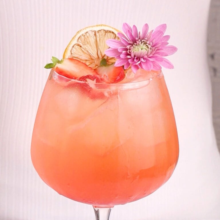 A pink iced mocktail in a stemmed glass, garnished with a dried lemon slice, strawberry slices, and a purple flower, set against a light background.