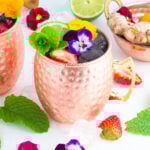 A copper mug filled with a spring moscow mule mocktail, garnished with edible flowers, mint leaves, lime, and strawberries. Ginger root, strawberries, mint, lime, and more flowers are arranged on a white surface around the mug.
