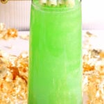 A tall glass filled with a bright green, St Patrick's Day mocktail topped with a gold stick holding sugar-coated, pastel candy strips. Gold foil decorates the background, and the glass has a gold geometric base.