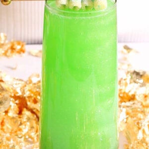 A tall glass filled with a bright green, St Patrick's Day mocktail topped with a gold stick holding sugar-coated, pastel candy strips. Gold foil decorates the background, and the glass has a gold geometric base.