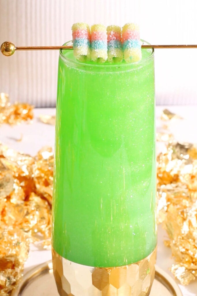 A tall glass filled with a bright green, St Patrick's Day mocktail topped with a gold stick holding sugar-coated, pastel candy strips. Gold foil decorates the background, and the glass has a gold geometric base.