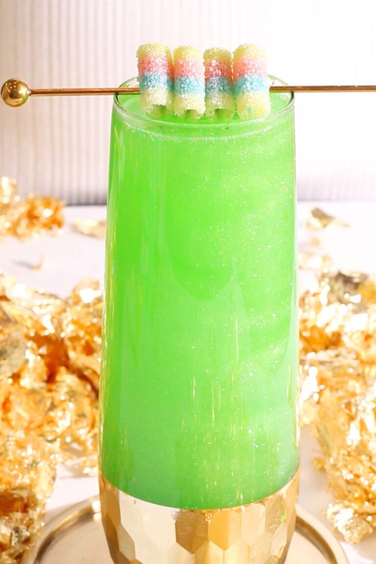 A tall glass filled with a bright green, St Patrick's Day mocktail topped with a gold stick holding sugar-coated, pastel candy strips. Gold foil decorates the background, and the glass has a gold geometric base.