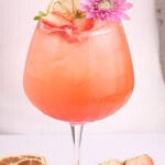 A pink cocktail in a large glass with ice, garnished with a dried lemon slice, fresh strawberry slices, and a purple flower. More dried lemon and strawberries are on the white surface nearby.