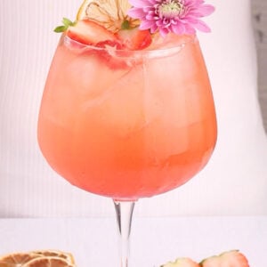 A pink cocktail in a large glass with ice, garnished with a dried lemon slice, fresh strawberry slices, and a purple flower. More dried lemon and strawberries are on the white surface nearby.