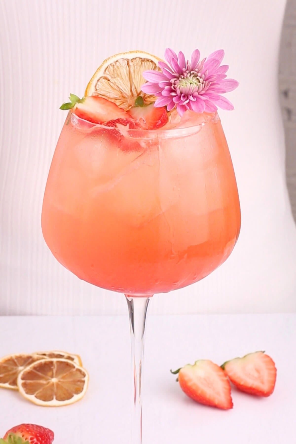 A pink cocktail in a large glass with ice, garnished with a dried lemon slice, fresh strawberry slices, and a purple flower. More dried lemon and strawberries are on the white surface nearby.