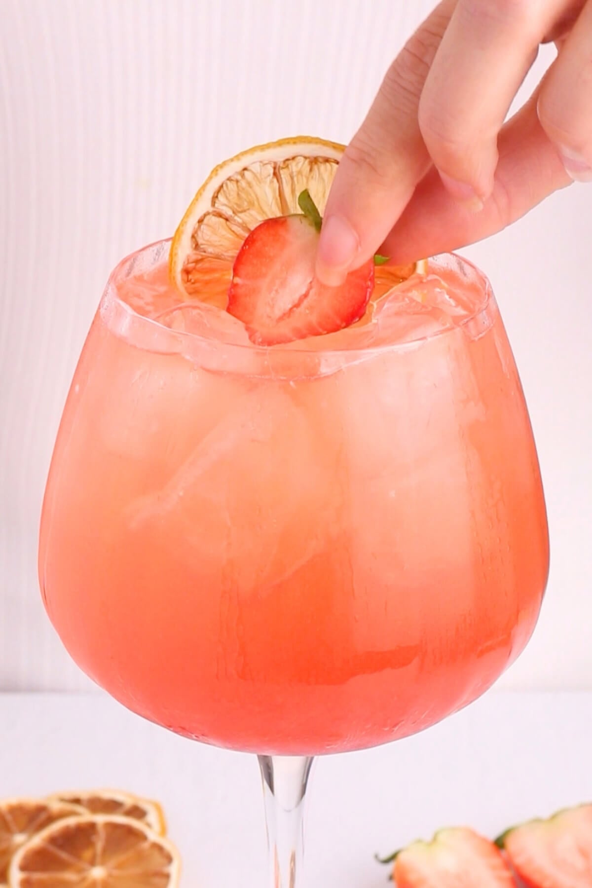A hand places a fresh strawberry and a dried citrus slice as garnish on top of a large, iced pink cocktail in a wide glass. Slices of citrus and strawberry are visible on the white surface below.