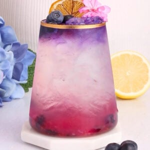 A colorful layered drink in a glass, transitioning from pink at the bottom to purple at the top, garnished with a dried orange slice, a blueberry, and a pink flower. Blueberries, a lemon half, and blue flowers are nearby.