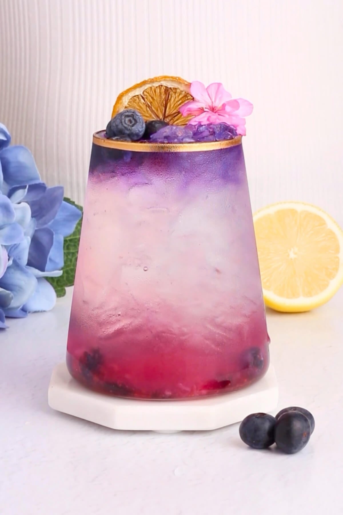 A colorful layered drink in a glass, transitioning from pink at the bottom to purple at the top, garnished with a dried orange slice, a blueberry, and a pink flower. Blueberries, a lemon half, and blue flowers are nearby.