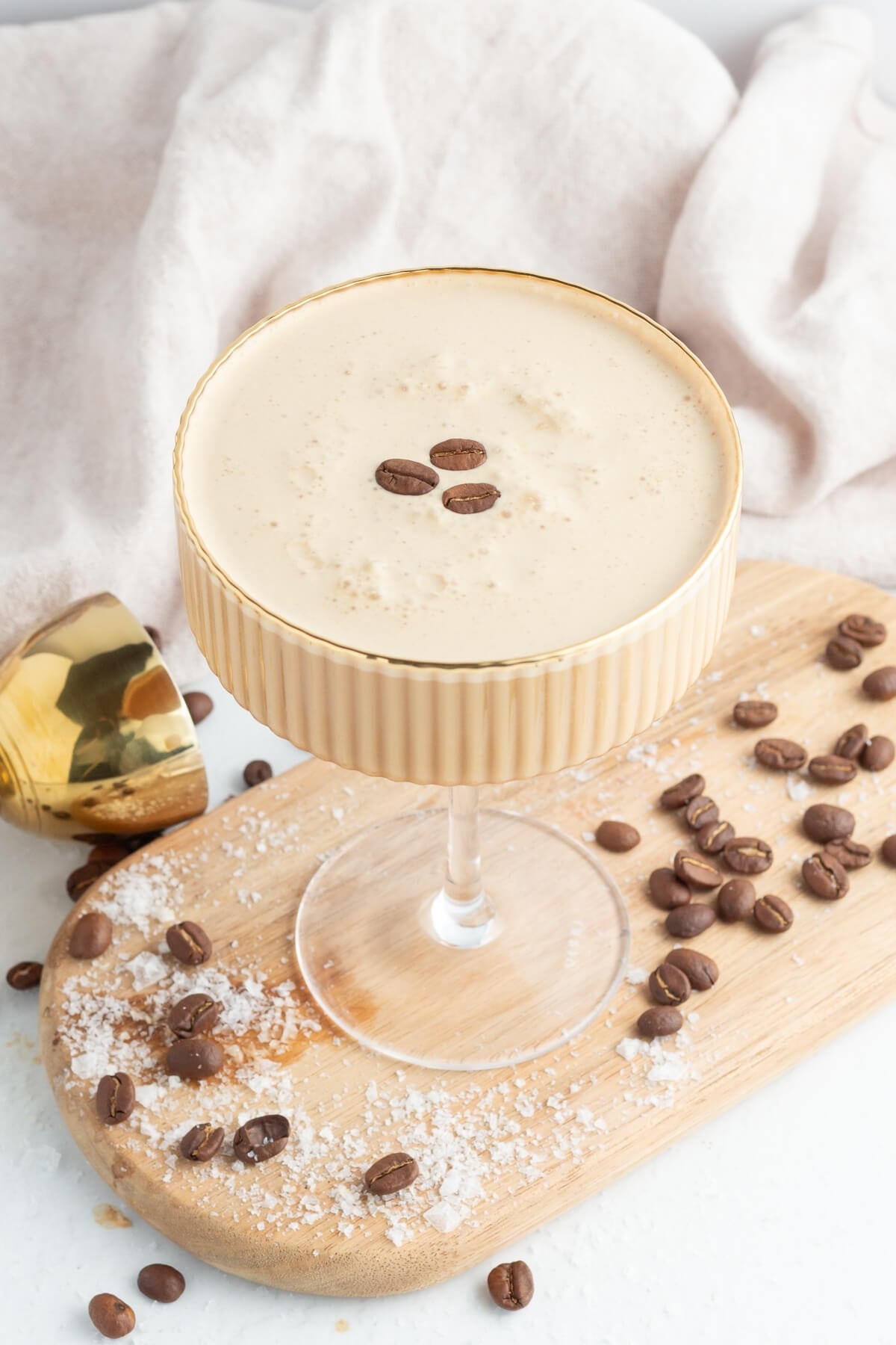 A creamy espresso martini garnished with three coffee beans sits in a gold-rimmed glass on a wooden board, surrounded by scattered coffee beans and a dusting of sugar, with a gold jigger nearby.