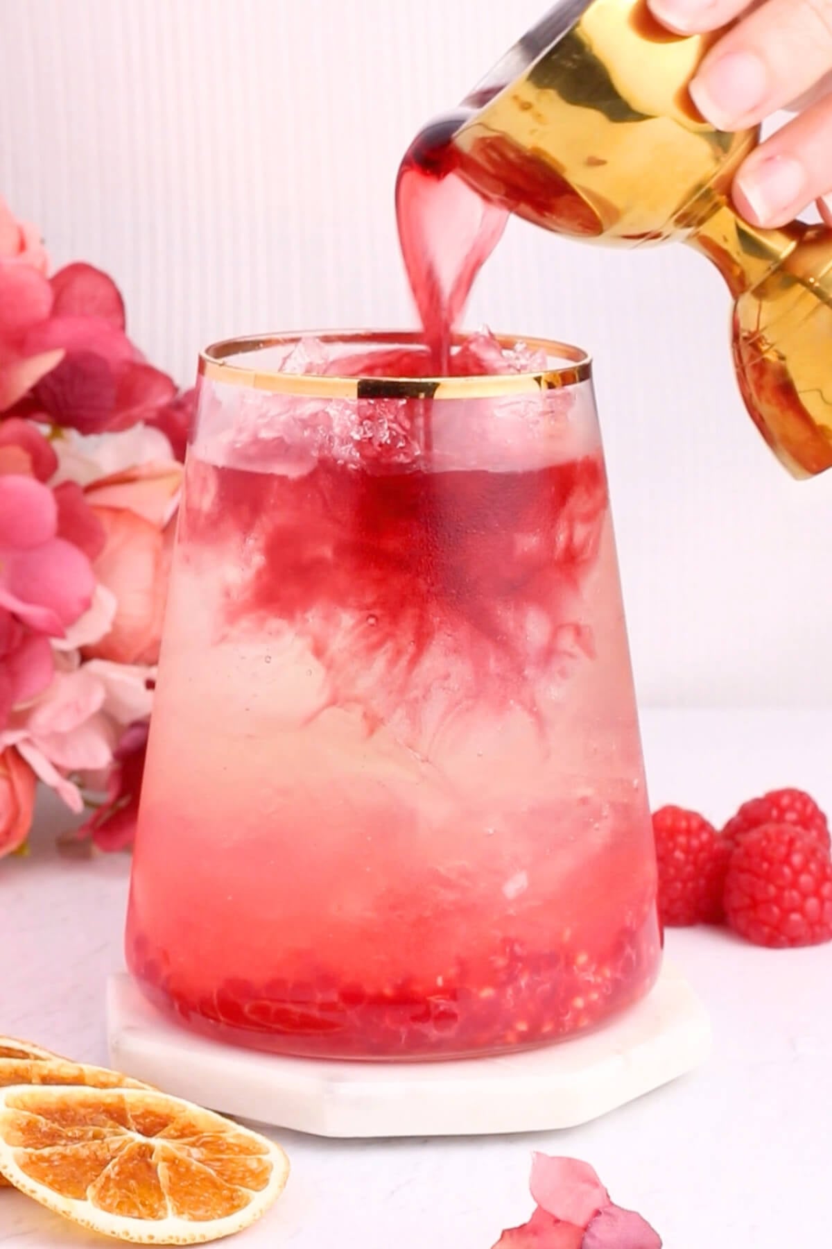 A hand pours vibrant red syrup into a glass of iced drink, creating swirling patterns. The glass is garnished with gold trim and surrounded by raspberries, dried orange slices, and pink flowers.