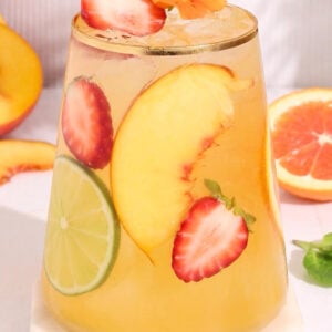 A glass of iced drink garnished with a peach slice, lime, strawberry slices, and orange flowers, surrounded by fresh fruit slices on a white surface.