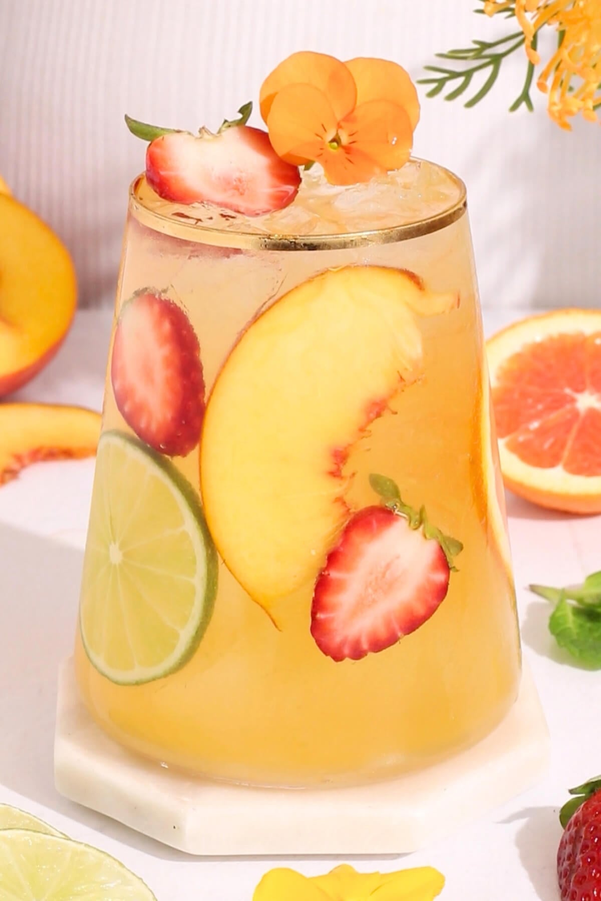 A glass of iced drink garnished with a peach slice, lime, strawberry slices, and orange flowers, surrounded by fresh fruit slices on a white surface.