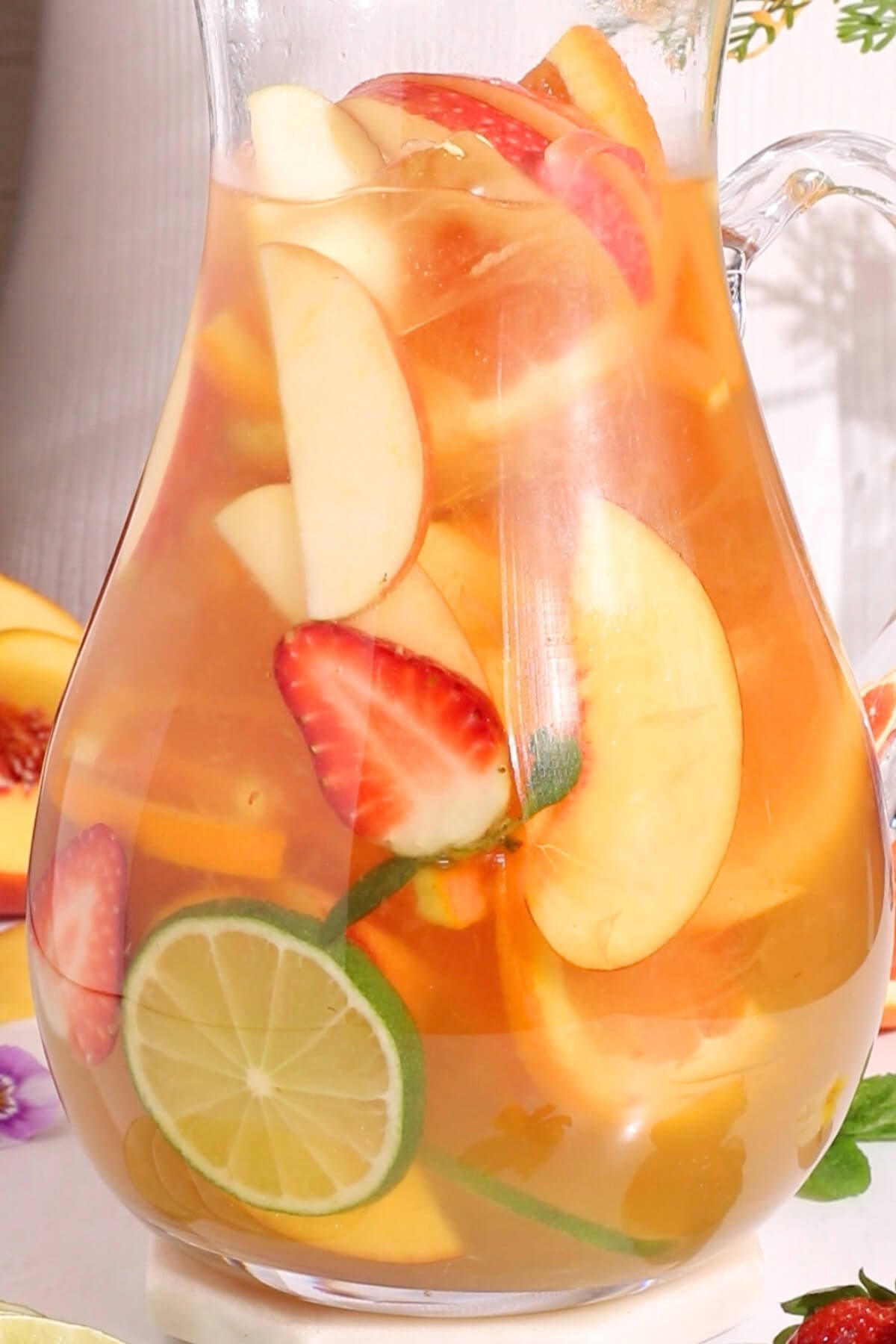 A clear glass pitcher filled with a light-colored beverage and slices of strawberries, apples, lime, and other assorted fruits. The pitcher is set on a bright surface with fruit and flowers around it.