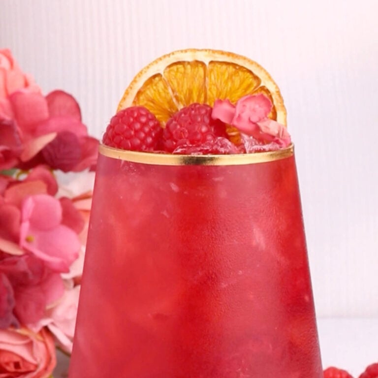 A vibrant red drink in a tall glass with gold rim, garnished with fresh raspberries, a dried orange slice, and ice. Pink flowers and raspberries are visible beside the glass.