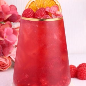 A vibrant red drink in a clear glass, garnished with raspberries, dried orange slices, and pink flower petals, sits on a white coaster. Pink flowers and raspberries are arranged beside the glass.