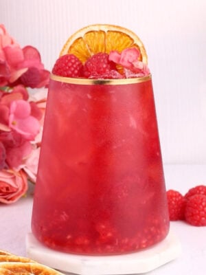 A vibrant red drink in a clear glass, garnished with raspberries, dried orange slices, and pink flower petals, sits on a white coaster. Pink flowers and raspberries are arranged beside the glass.