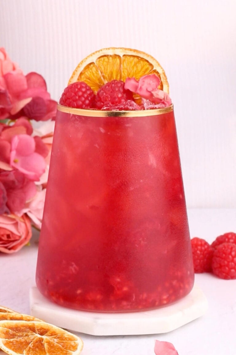 A vibrant red drink in a clear glass, garnished with raspberries, dried orange slices, and pink flower petals, sits on a white coaster. Pink flowers and raspberries are arranged beside the glass.
