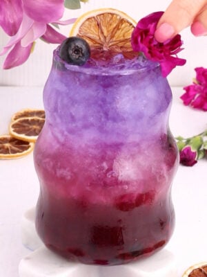 A vibrant layered drink transitions from deep purple at the bottom to pale lavender at the top, garnished with a dried orange slice, blueberry, and edible flower, with a hand placing a pink blossom on the rim. Pink flowers and dried oranges surround the glass.