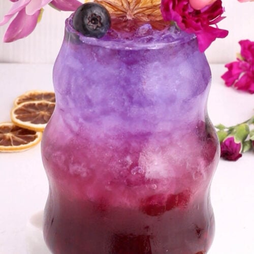 A vibrant layered drink transitions from deep purple at the bottom to pale lavender at the top, garnished with a dried orange slice, blueberry, and edible flower, with a hand placing a pink blossom on the rim. Pink flowers and dried oranges surround the glass.