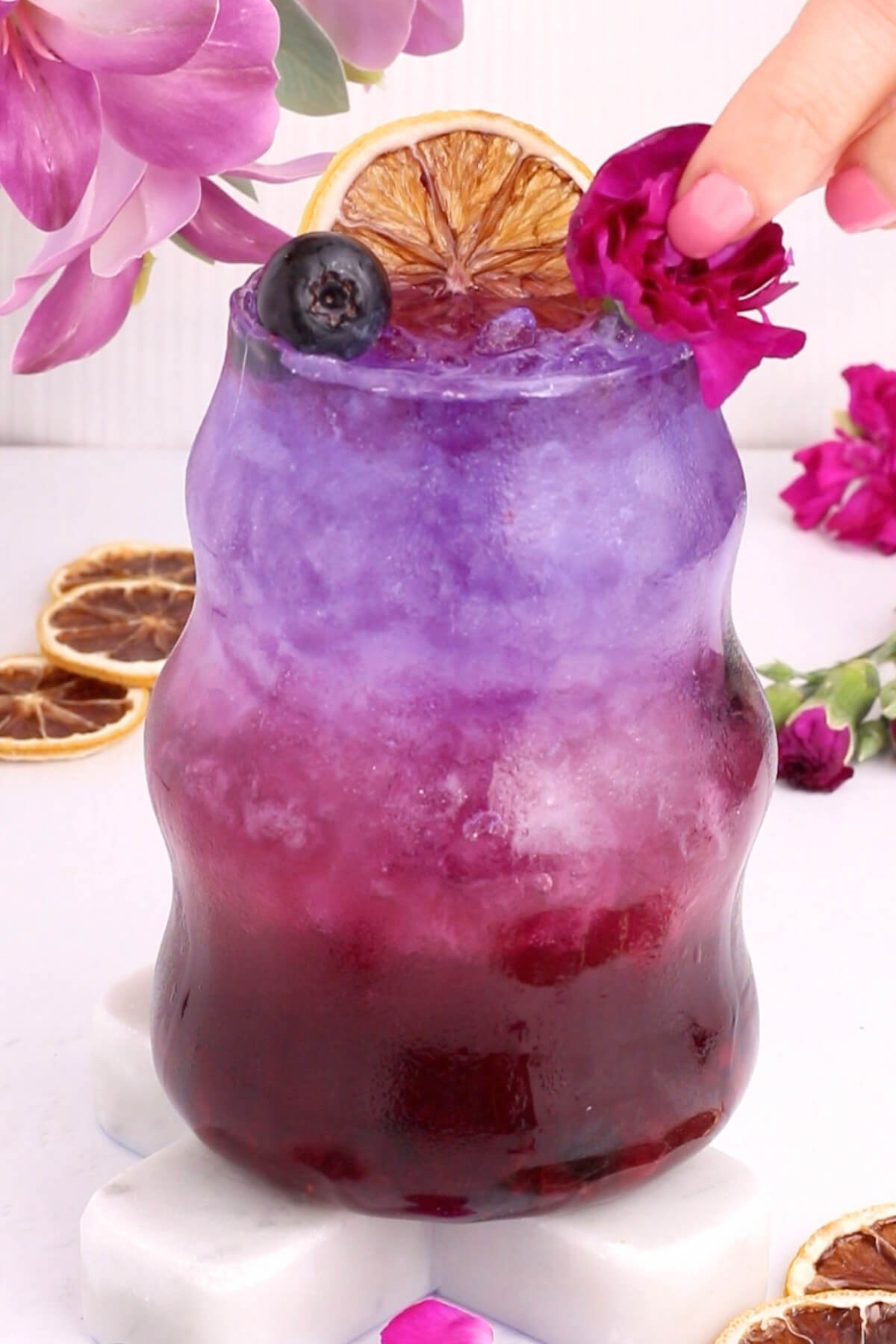 A vibrant layered drink transitions from deep purple at the bottom to pale lavender at the top, garnished with a dried orange slice, blueberry, and edible flower, with a hand placing a pink blossom on the rim. Pink flowers and dried oranges surround the glass.