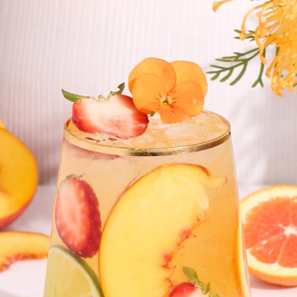 A glass of iced fruit drink garnished with peach slices, strawberries, lime, and an orange edible flower, with more fruit and flowers in the background.