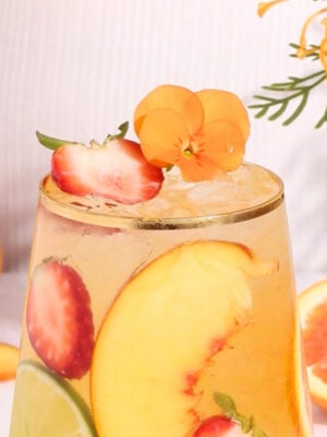 A glass of iced fruit drink garnished with peach slices, strawberries, lime, and an orange edible flower, with more fruit and flowers in the background.