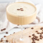 A creamy coffee cocktail garnished with three coffee beans, served in a ribbed glass on a wooden board, with scattered coffee beans and a light cloth in the background.