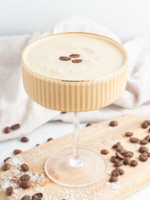 A creamy coffee cocktail garnished with three coffee beans, served in a ribbed glass on a wooden board, with scattered coffee beans and a light cloth in the background.