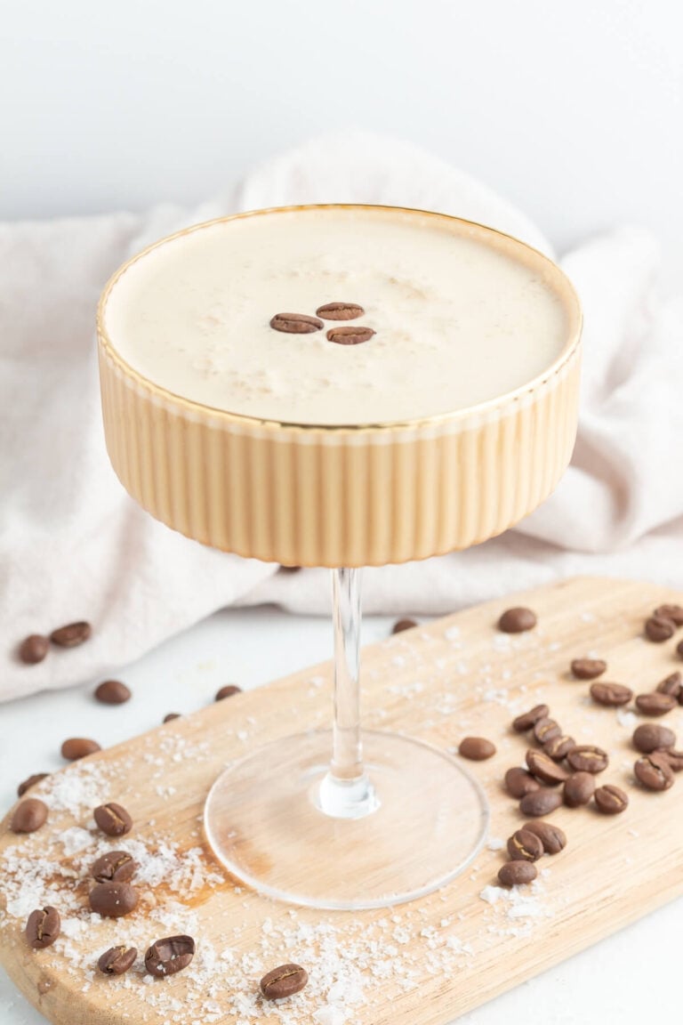 A creamy coffee cocktail garnished with three coffee beans, served in a ribbed glass on a wooden board, with scattered coffee beans and a light cloth in the background.