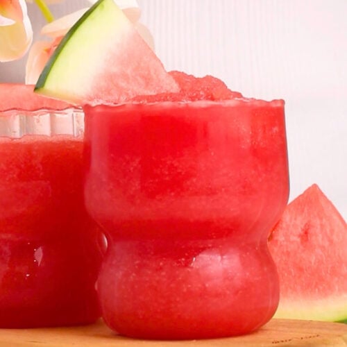 A vibrant red watermelon slushie in a rounded glass sits on a wooden board, garnished with a fresh watermelon slice. Another glass and additional watermelon slices are in the background, along with soft pink flowers.