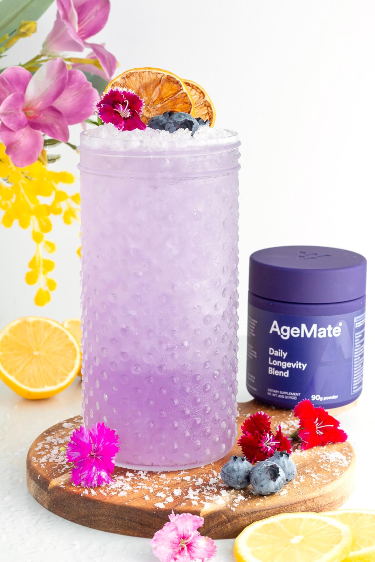 A tall, textured glass filled with a purple drink topped with crushed ice, dried citrus, blueberries, and edible flowers. AgeMate supplement container, flowers, lemon slices, and blueberries surround the glass on a wooden board.