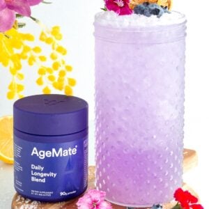 A glass of purple drink garnished with blueberries, edible flowers, and dried citrus slices sits next to a purple AgeMate Daily Longevity Blend container on a wooden board with flowers, lemon slices, and blueberries around.