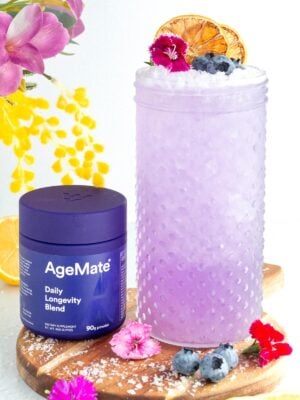 A glass of purple drink garnished with blueberries, edible flowers, and dried citrus slices sits next to a purple AgeMate Daily Longevity Blend container on a wooden board with flowers, lemon slices, and blueberries around.